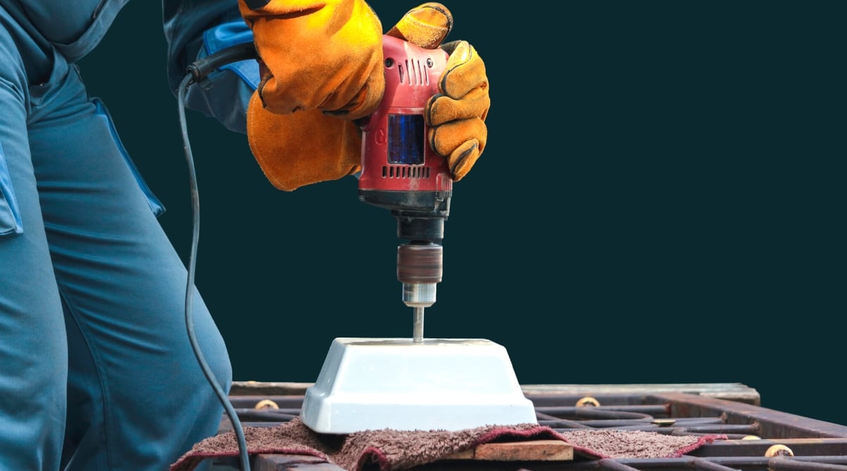 Close-up of male hands drilling a drainage hole in a small square ceramic pot, outdoors, against a blue wall. The drill is large, red. The man is wearing orange gardening gloves and blue overalls.