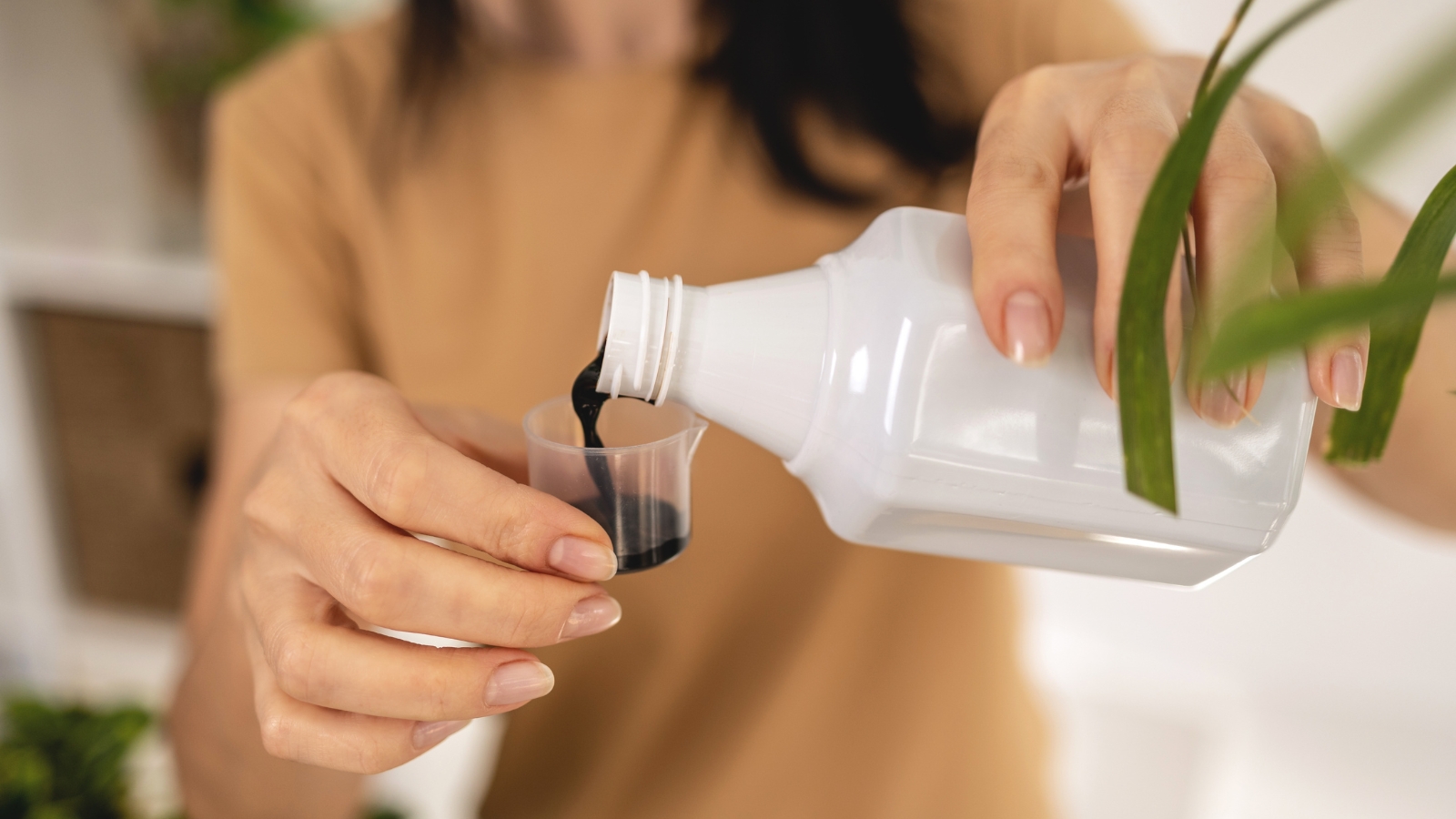 Close up of a woman in a tan shirt, pouring a dark liquid into a small plastic cap out of a white plastic container.